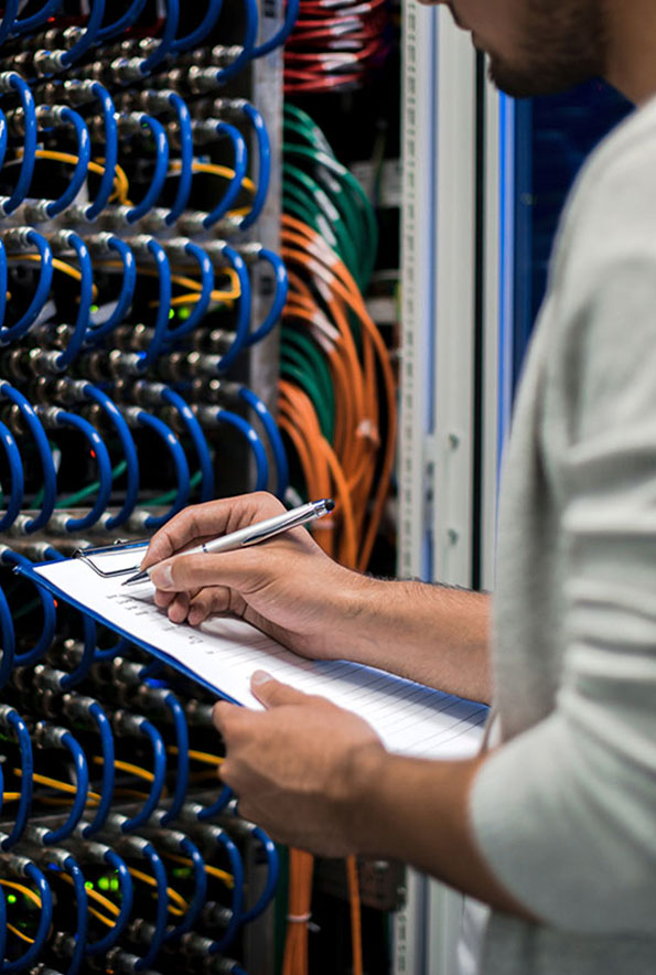 Person holding a clipboard and pen, checking or recording information in front of a server rack with organised blue and orange network cables.