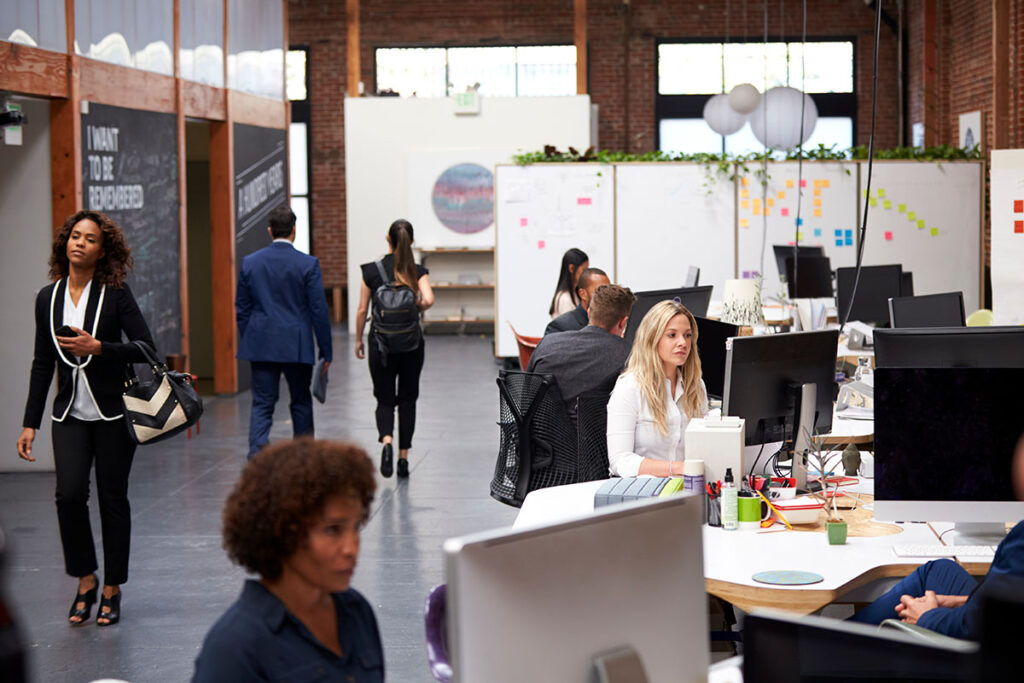 Modern open-plan office in Huddersfield with people working on computers at desks and others walking; exposed brick walls, large windows, and whiteboards in the background.