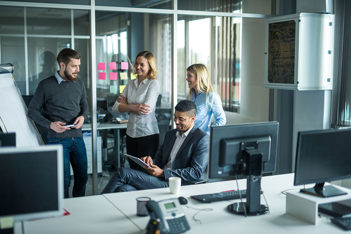 Four colleagues in business attire have a discussion near a flipchart in a modern office with computers and glass walls. One person is seated, looking at a tablet.