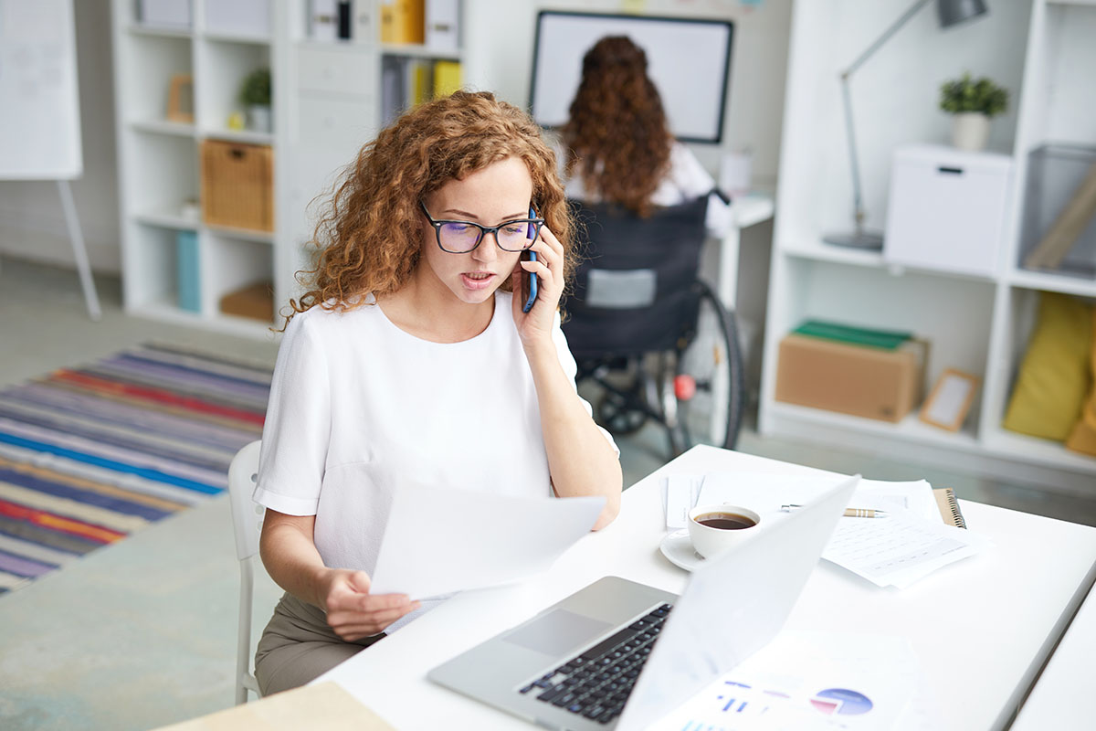 Woman with curly hair and glasses talks on the phone while looking at documents at a desk; another person in a wheelchair works at a computer in the background.